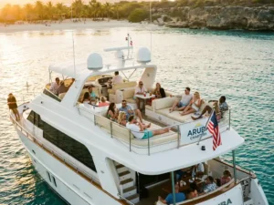 White charter boat with diverse passengers on multiple decks cruising turquoise waters off Aruba's palm-lined coast at sunset.