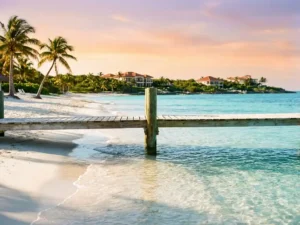 Turquoise Caribbean waters with white sand beach, wooden dock, palm trees, and luxury resort at golden hour